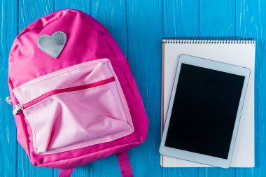 mavi ahşap arka plan üzerinde boş ekran, boş ders kitabı ve pembe sırt çantası ile dijital tablet üstten görünüm 