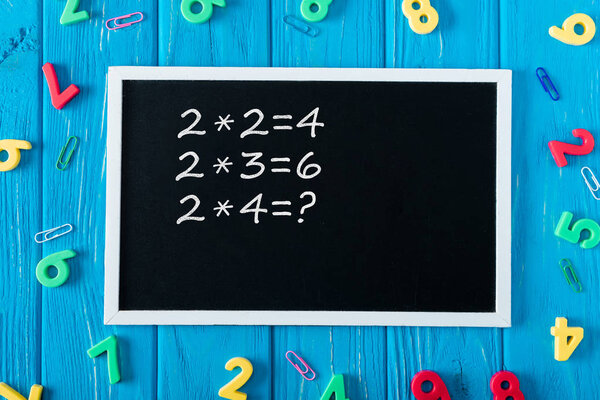 top view of blackboard with multiplication table, colorful numbers and paper clips on blue wooden background 