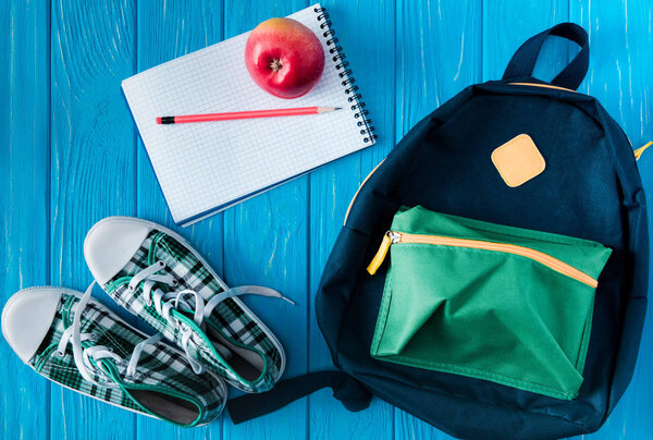 top view of sneakers, rucksack, empty notebook, apple and pencil on blue wooden background