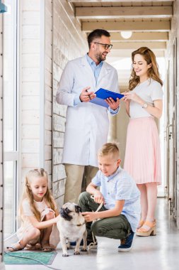 veterinarian showing something in clipboard to woman, kids playing with pug dog at veterinary clinic  clipart