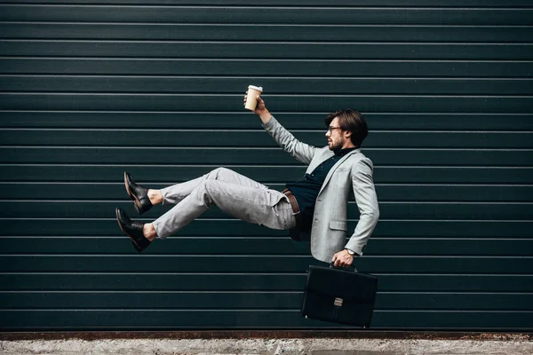 side view of young businessman falling with paper cup of coffee in front of roller gate