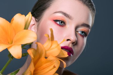 close-up portrait of attractive young woman with fashionable makeup and orange lilium flowers looking away isolated on grey clipart
