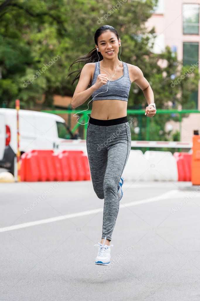 smiling young asian female jogger running in earphones running at city street