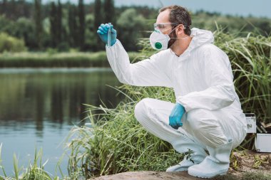 side view of male scientist in protective suit examining sample of water in test flask outdoors  clipart