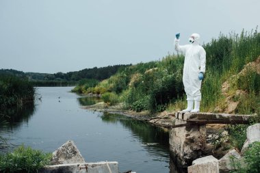 male scientist in protective mask and suit looking at sample of water in test flask outdoors  clipart