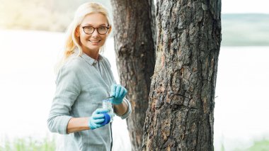 smiling female scientist in eyeglasses putting sample by tweezers in jar near trees outdoors  clipart