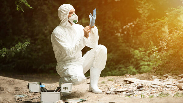 selective focus of male scientist in protective mask and suit putting on latex glove in forest