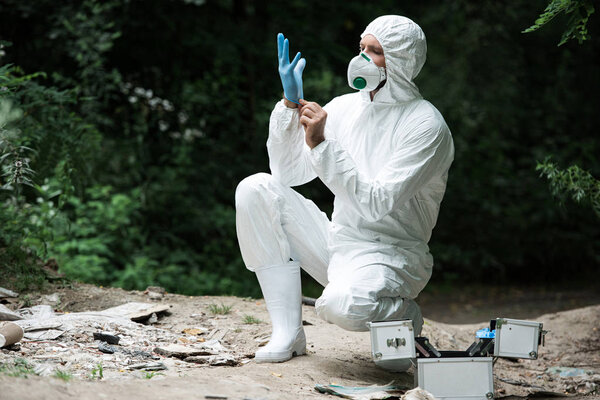 male scientist in protective mask and suit putting on latex glove in forest