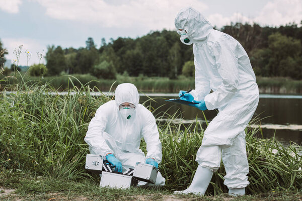 male scientist in protective mask and suit opening working suitcase while his female colleague writing in clipboard 