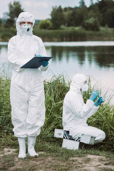 Wissenschaftlerin Schutzmaske Und Anzug Schreibt Klemmbrett Während Ihr Kollege Wasserproben — Stockfoto