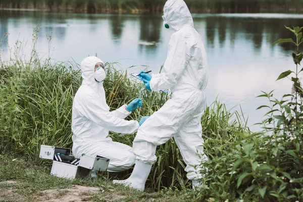 Wissenschaftlerin Schutzmaske Und Anzug Schreibt Klemmbrett Während Ihr Kollege Eine — Stockfoto