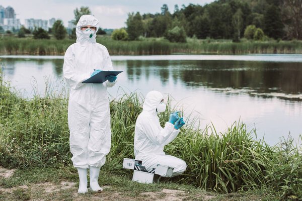 selective focus of female scientist in protective mask and suit writing in clipboard while her colleague putting sample of water near river outdoors 