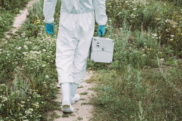 обрезанное изображение мужчины-ученого в защитном костюме, идущего с рабочим чемоданом на лугу
