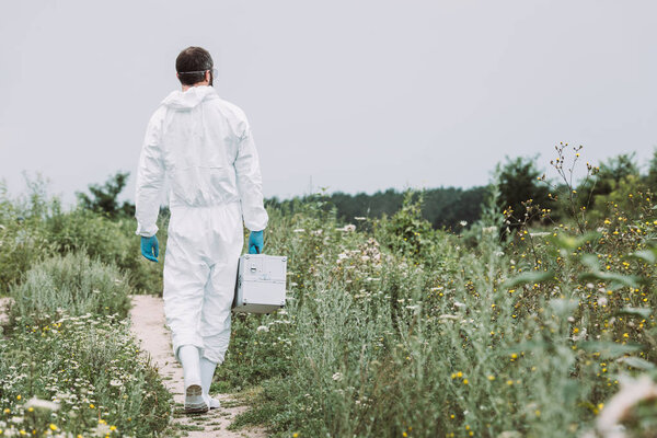 rear view of male scientist in protective suit walking with working suitcase in meadow 