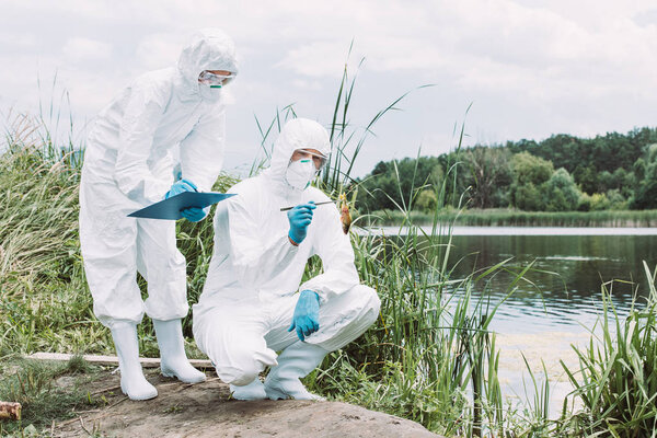 two scientists in protective masks and suits examining fish and writing in clipboard near river 