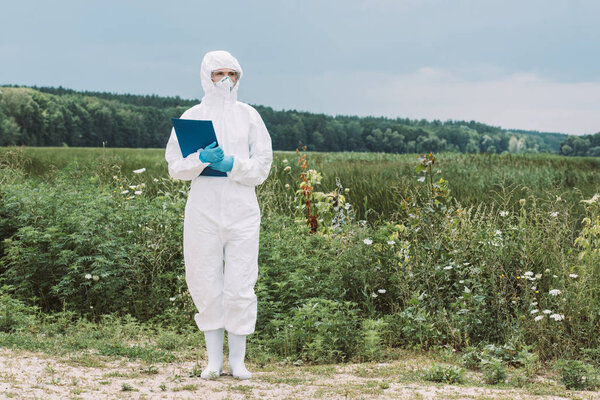 female scientist in protective suit and googles holding clipboard in meadow 