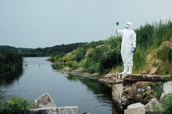 male scientist in protective mask and suit looking at sample of water in test flask outdoors 