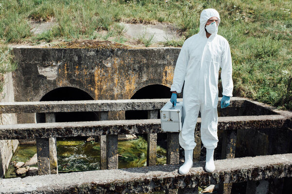 male scientist in protective mask and suit holding working suitcase near sewerage 