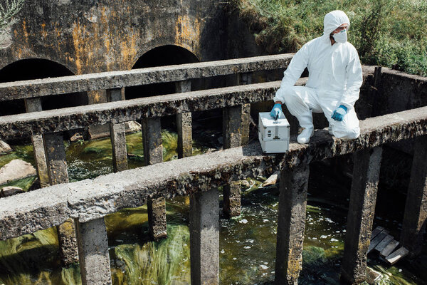 male scientist in protective mask and suit sitting with working suitcase near sewerage 