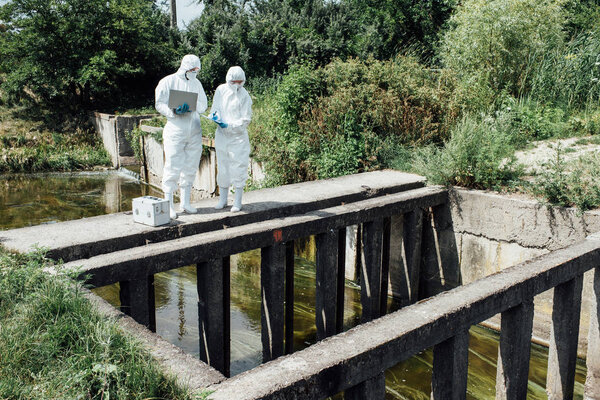 two scientists in protective suits and masks working with laptop near sewerage 