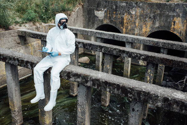 male scientist in protective mask and suit writing in clipboard while sitting near sewerage 