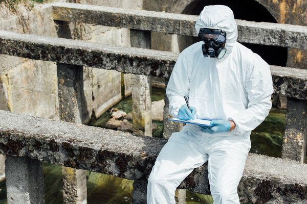 male scientist in protective mask and suit writing in clipboard while sitting near sewerage 