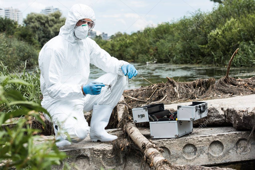 male scientist in protective suit holding sample of soil by tweezers and water in test flask outdoors 