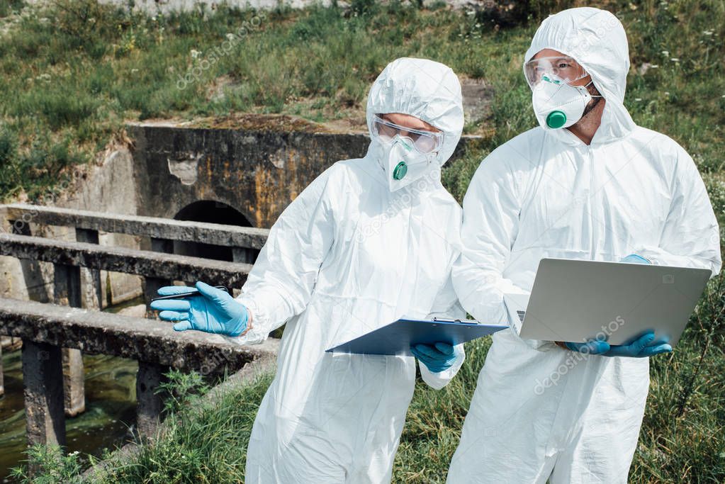 selective focus of female scientist with clipboard pointing to male colleague with laptop near sewerage 