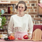 Beautiful adult housewife in vintage clothes looking at camera at kitchen
