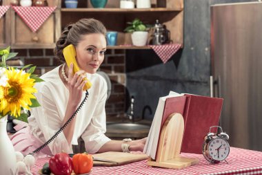 Retro kablolu telefonla konuşurken ve mutfak kameraya bakarak mutlu yetişkin ev hanımı