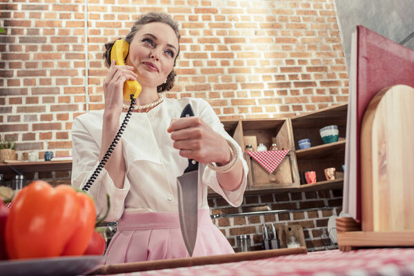 вид снизу на улыбающуюся взрослую домохозяйку с ножом разговаривающую с помощью желтого проводного телефона на кухне
