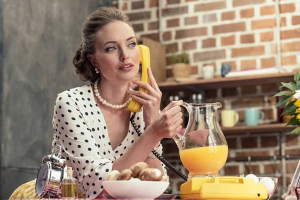 キッチン ヴィンテージ有線電話で話しているオレンジ ジュースのジョッキを持って魅力的なアダルト主婦 — ストック写真