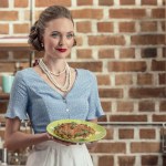 Smiling adult housewife in vintage clothes holding plate with mushroom cake at kitchen