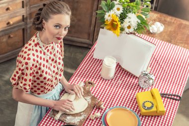 oklava ile hamur yoğurma ve mutfak kameraya bakarak güzel yetişkin ev hanımı yüksek açılı görünüş