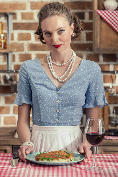 smiling adult housewife with glass of red wine and cake looking at camera at kitchen