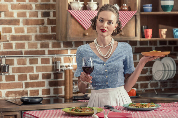attractive adult housewife with glass of red wine and freshly baked cake looking at camera at kitchen