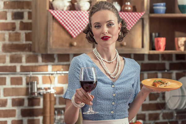 beautiful adult housewife with glass of red wine and freshly baked cake looking at camera at kitchen