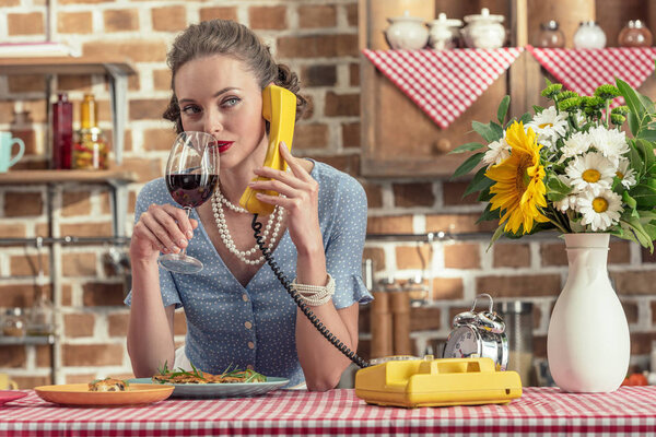 beautiful adult housewife with glass of wine talking by vintage rotary phone at kitchen