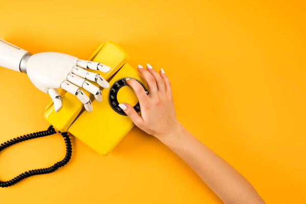 cropped shot of woman and robot reaching for vintage phone on yellow
