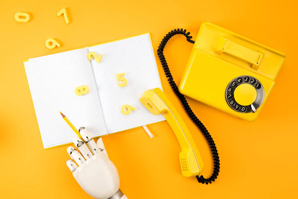 обрезанный снимок роботизированной руки, записанной чистым блокнотом на желтом столе с винтажным телефоном и математическими номерами
