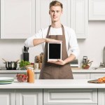 Guapo joven sosteniendo la tableta con pantalla en blanco en la cocina
