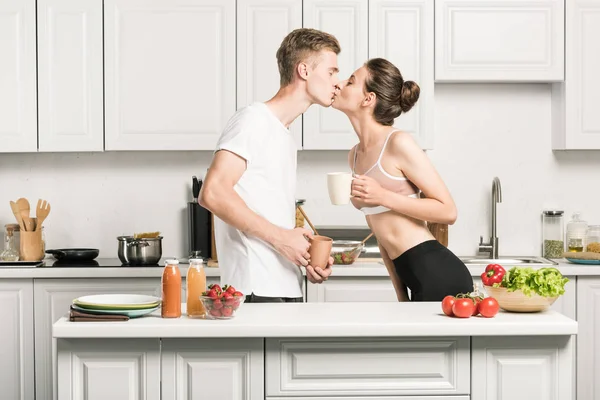 年轻夫妇在厨房里抱着杯子亲吻的侧面视图 — 图库照片