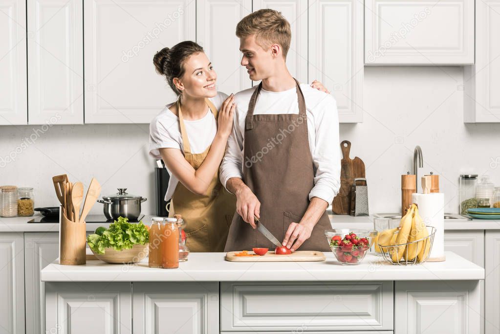 girlfriend hugging boyfriend while he cutting tomatoes for salad in kitchen