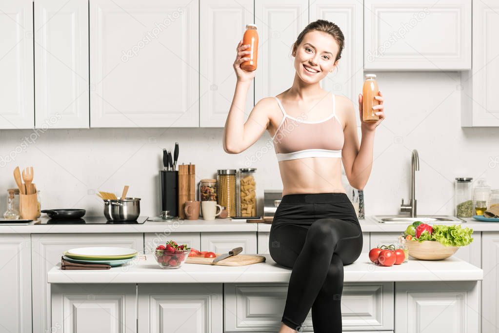 happy attractive girl sitting on kitchen counter and showing healthy juice in bottles