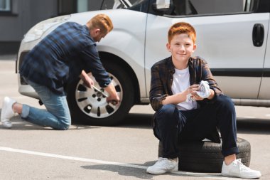 father changing tire in car with wheel wrench, son sitting and looking at camera clipart