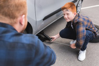 son lifting car with floor jack for changing tire and looking at father clipart