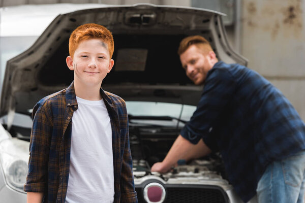 father repairing car with open hood, smiling son looking at camera