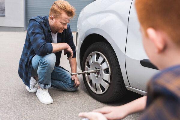 father changing tire in car with wheel wrench, son sitting near