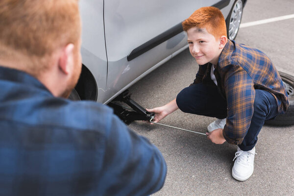 son lifting car with floor jack for changing tire and looking at father
