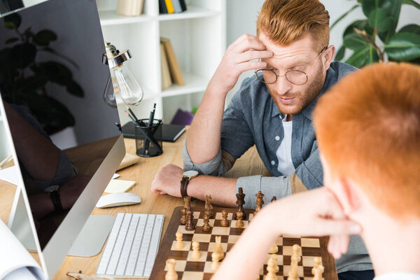 pensive red hair father and son playing chess at home